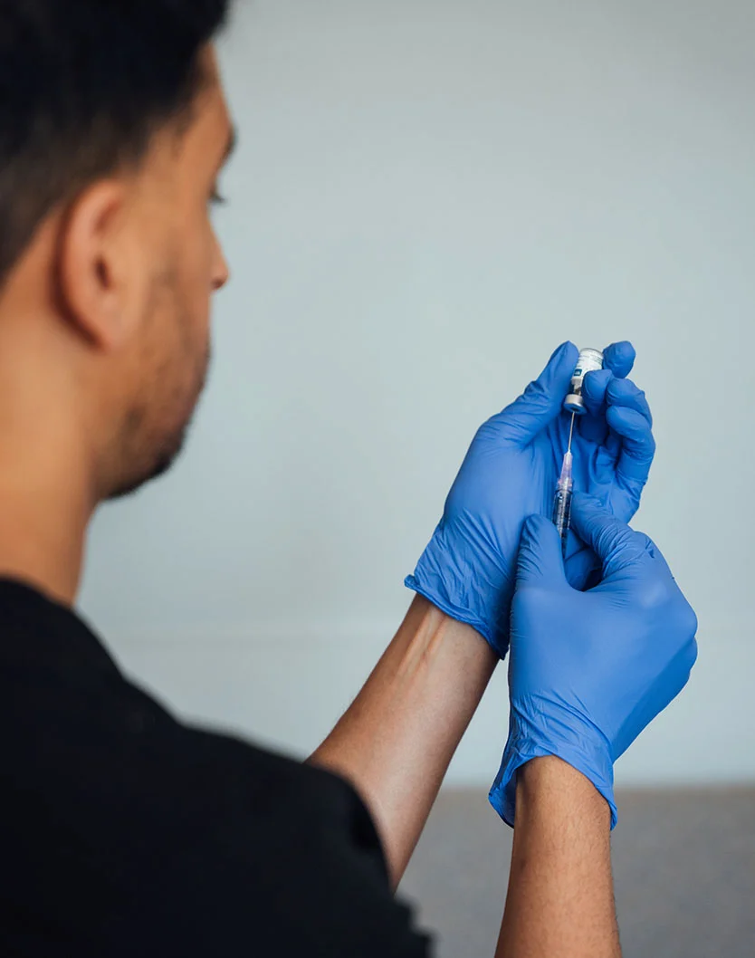 Close-up of a medical professional in dark clothing and blue nitrile gloves preparing to administer an injection. They are focused on the task, holding a syringe and small vial with precision. The light gray-white background suggests a clinical setting. - Botox and Wrinkle Relaxers in Glenview, IL