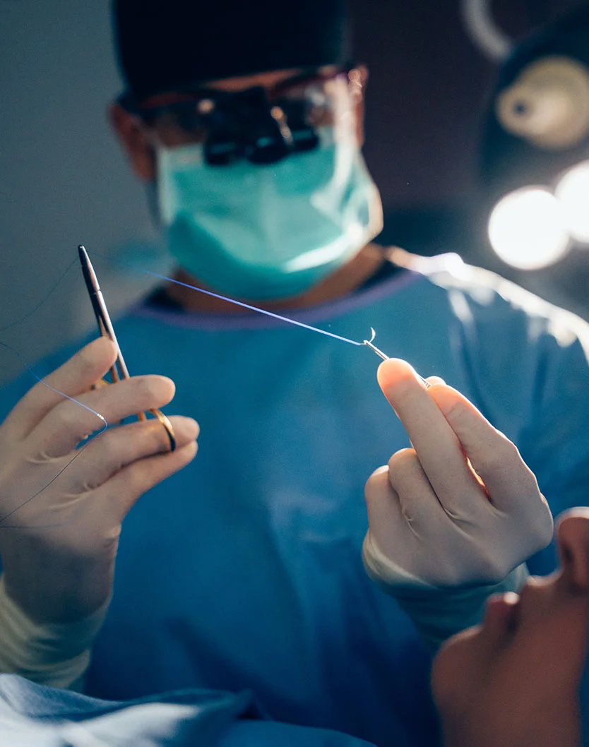 Close-up of Dr. Shastri performing a procedure. Wearing a surgical mask, glasses, and blue scrubs, he is focused on handling surgical instruments and suture material. The patient's face and part of their body are visible, with surgical equipment and lighting in the blurred background, emphasizing the ongoing procedure. - Buccal Fat Removal in Glenview, IL
