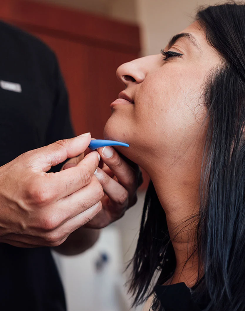 Close-up of a person receiving a facial treatment. A light-blue tool is applied near their chin, with the hands of a professional visible. The background is blurred with a reddish-brown hue, and the lighting highlights the person's face. - Chin Augmentation in Glenview, IL