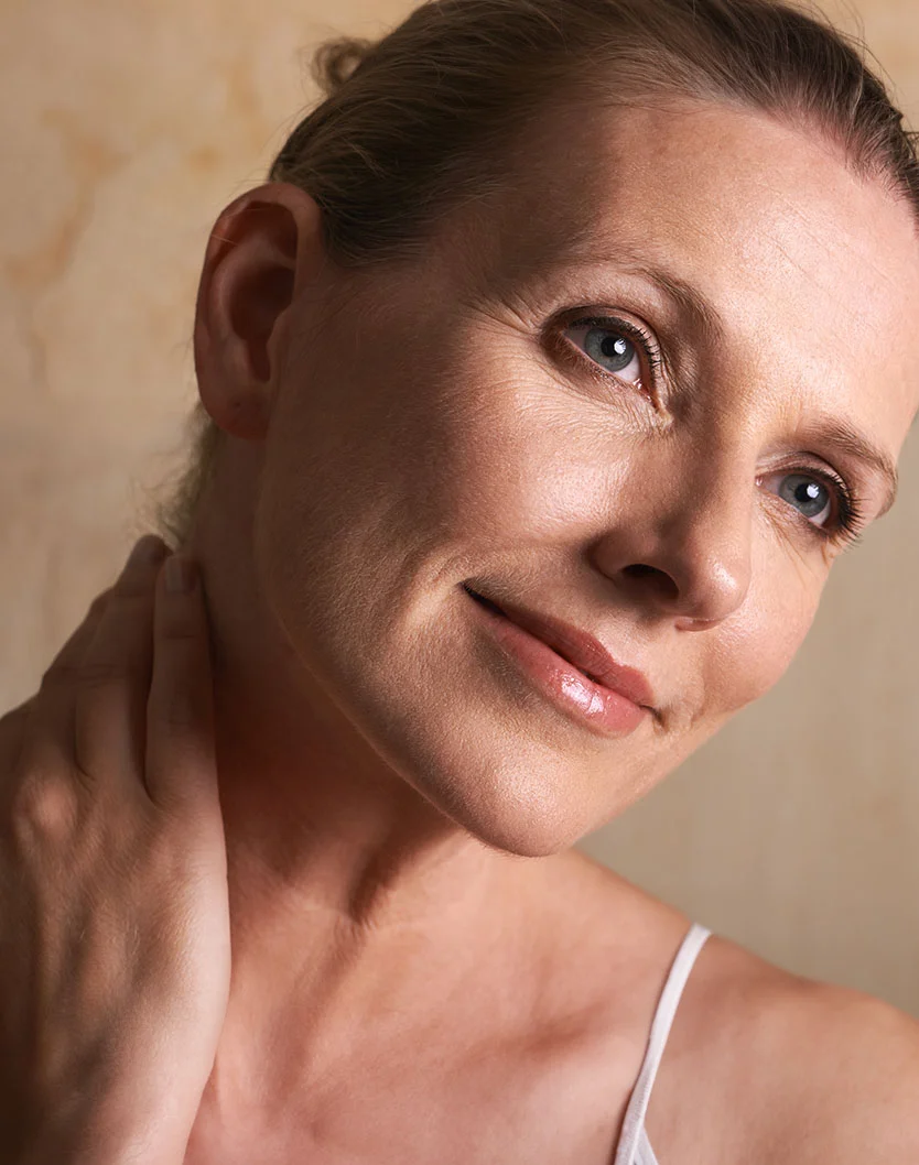Close-up of a middle-aged woman with light blonde hair in a bun. She has natural wrinkles, a relaxed expression, and a subtle smile. Her hand rests on her neck, with soft, diffused lighting and a neutral background. - Deep Plane Necklift in Glenview, IL