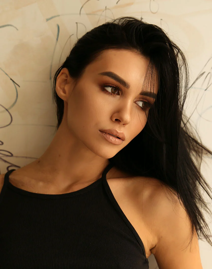 Close-up of a woman with long, dark hair styled in loose waves, wearing a black halter top. Her light-tan skin, brown eyes, and neutral makeup, including rosy-brown lipstick, create a pensive expression. The out-of-focus light beige background with abstract markings highlights her features. - Hairline Lowering in Glenview, IL
