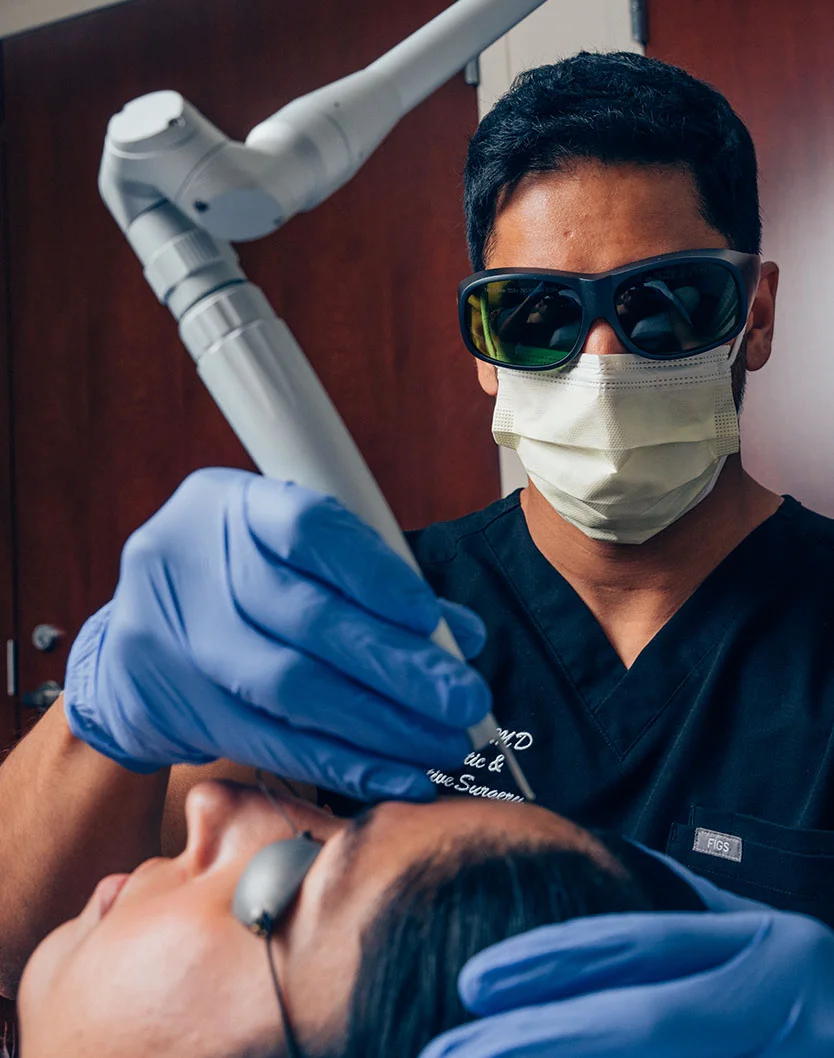 Extreme close-up of a dermatologist in dark scrubs and protective gear performing a procedure on a patient wearing blue nitrile gloves. A skin treatment device is near the patient’s face, with focused lighting and a sterile medical environment in the background. - Laser Services in Glenview, IL