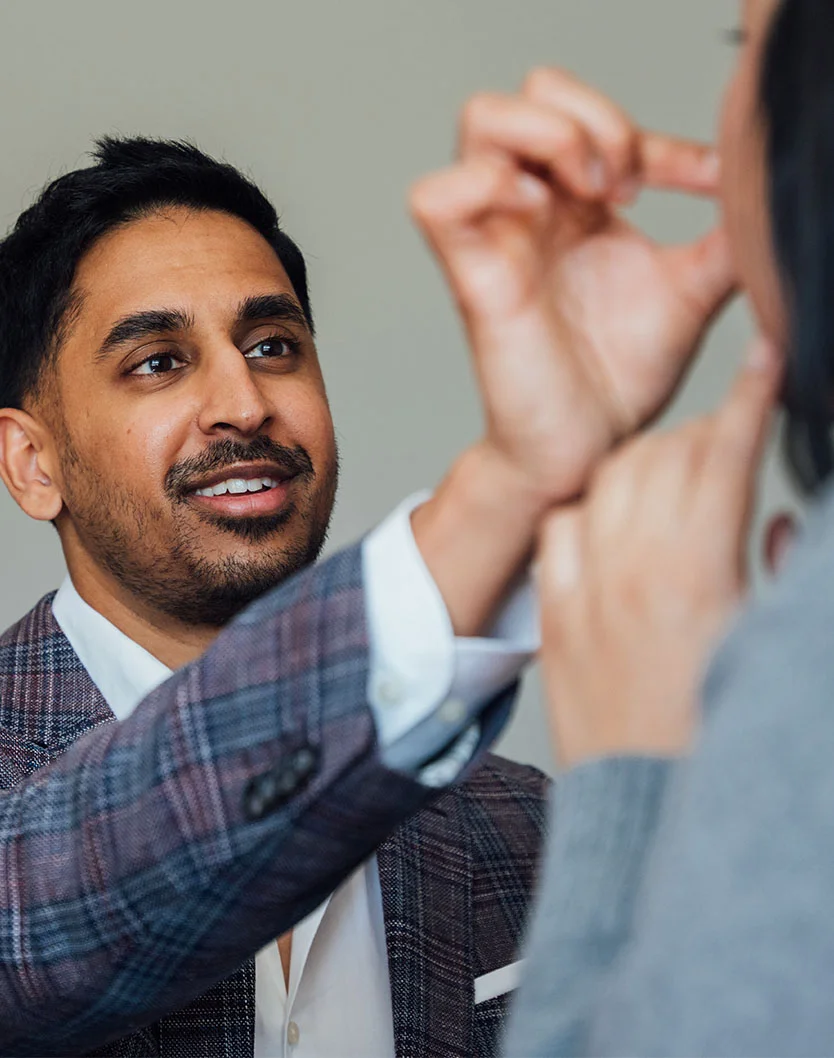 Close-up of Dr. Shastri in a consultation, gently touching a woman's face. He wears a patterned suit jacket, and she has dark hair in a light gray or blue top. The neutral gray background and soft lighting focus on their interaction, suggesting a medical or therapeutic discussion. - Lip Lift in Glenview, IL