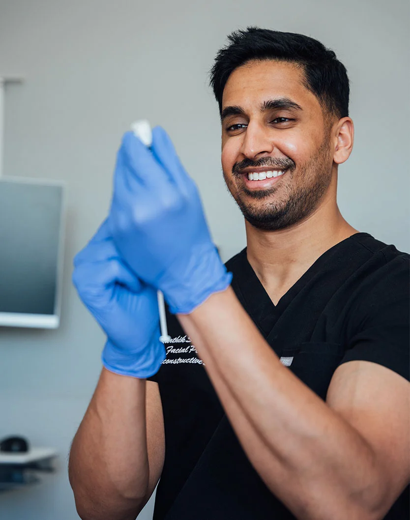 Close-up of Dr. Shastri smiling, wearing blue nitrile gloves and a dark scrub top. He holds a small medical instrument, with a monitor and blurred wall in the background. The lighting is soft, creating a clean, professional look. - Liquid Rhinoplasty in Glenview, IL