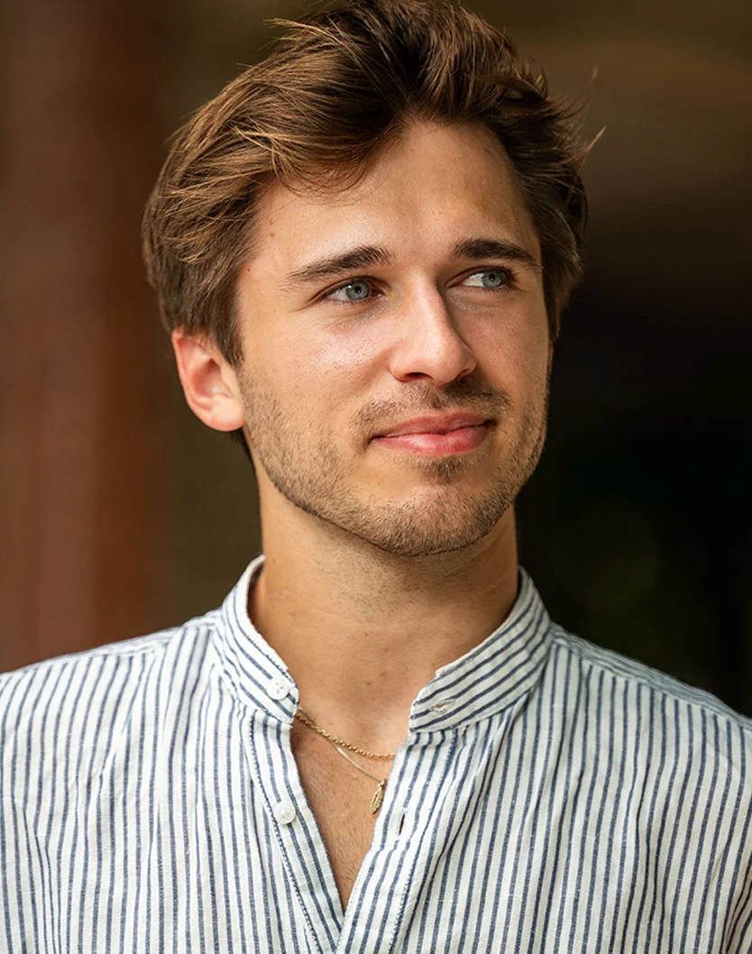 Close-up of a young man with light brown, tousled hair and light blue eyes. He has a neutral expression and light-toned skin, appearing in his late teens or early twenties. He wears a light-colored collared shirt with dark blue vertical stripes and a mandarin collar, with a small gold pendant necklace visible. The background is a blurred dark brownish-orange, focusing on his face and chest. - Otoplasty in Glenview, IL