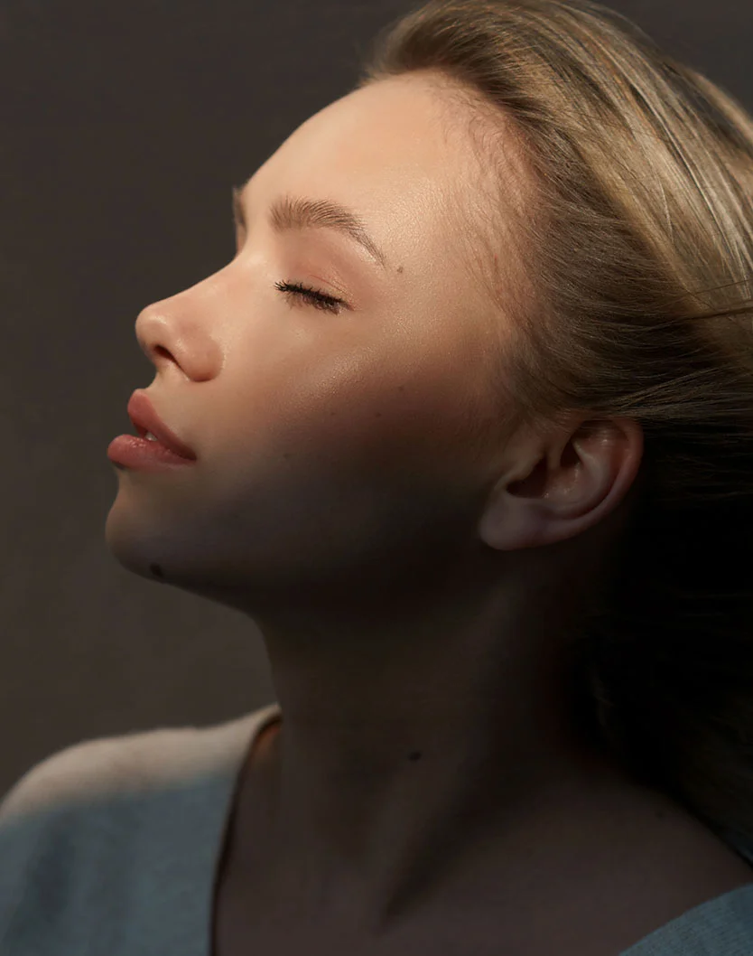 Close-up portrait of a young woman with light skin and long, light brown hair. Her eyes are closed, and her expression is serene. Soft, diffused lighting casts gentle shadows on her face, highlighting subtle makeup. She wears a light-colored top, partially visible at the bottom of the frame. The dark, neutral background enhances focus on her features. - Rhinoplasty in Glenview, IL