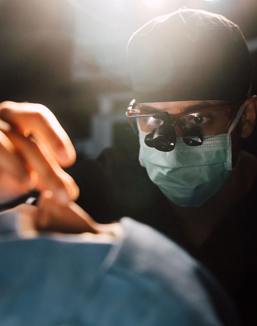 Close-up of Dr. Shastri in a surgical procedure, focused and wearing a mask and magnifying glasses. Their face and upper body are clearly visible, with dark scrubs and a sterile environment. In the background, hands and torso in blue-gray scrubs suggest a patient or another team member, emphasizing the precision of the delicate procedure. - Septoplasty in Glenview, IL